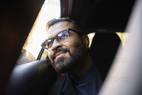 Indian businessman rides in a car on a sunny winter day, smiling warmly and looking slightly upwards throughout the window