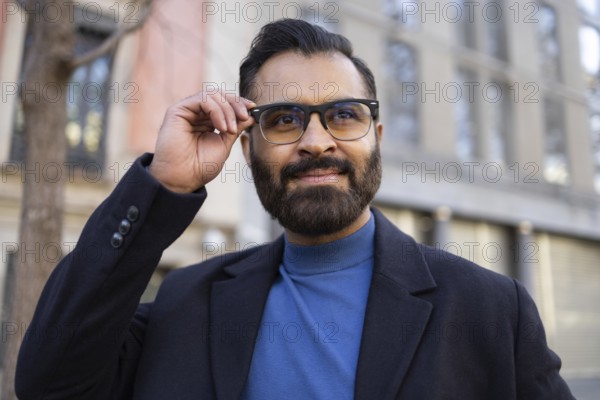 An Indian businessman in stylish winter attire, adjusting his glasses while smiling confidently outdoors, near an urban backdrop