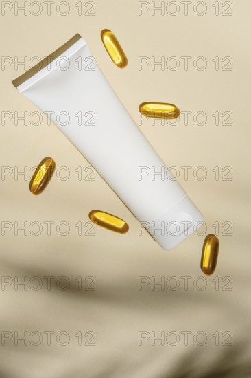 Top view of a blank white cosmetic tube floats amidst golden oil capsules, all set against a soft beige background, suggesting luxury skincare