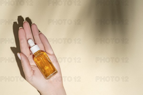 Top view of a cropped unrecognizable female hand holding a transparent dropper bottle filled with a golden cosmetic liquid, presented on a serene beige background with soft shadows