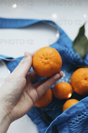Cropped unrecognizable image of a hand holding a tangerine over a blue string bag filled with tangerines, highlighting a moment of healthy lifestyle choice