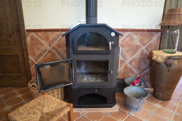 A traditional wood burning stove with its door open, situated in a cozy room corner showing warm, rustic decor including a woven basket, ceramic tiles, and wooden furniture