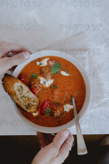 From above cropped unrecognizable person holding a bowl of tomato soup with meatballs, bread, and fresh basil