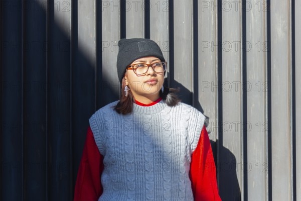 A Latina woman in stylish urban attire, including a knit vest and beanie, stands confidently by a modern, shadowed wall Her fashion sense blends casual with contemporary