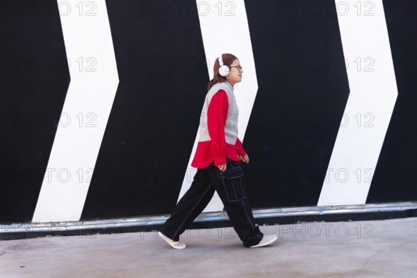Latina woman in a vibrant red shirt and headphones walks confidently against a bold black and white mural backdrop The stylish urban scene captures youthful energy and diversity