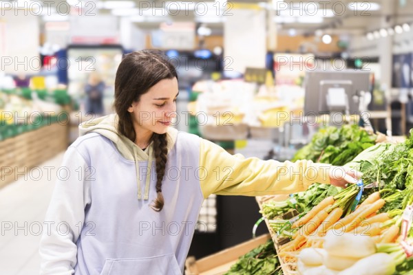 A young woman with a braid, wearing a casual hoodie, selects fresh vegetables in a brightly lighted supermarket aisle. She is looking down at the products