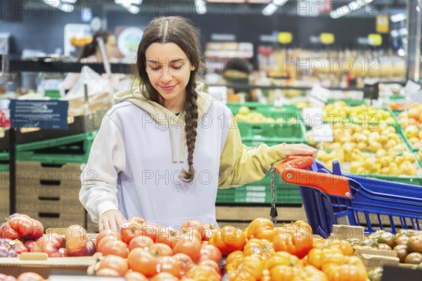 A happy young woman picking tomatoes from a stall in a supermarket, examining them carefully while holding a shopping basket