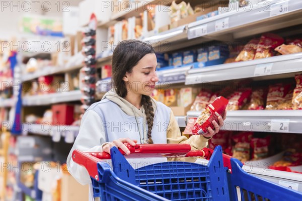 A woman with a braid examines a product in a supermarket aisle, pushing a shopping cart