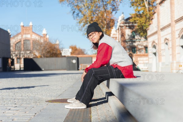 A young Latina woman in a red shirt and black pants sits on a concrete bench in an urban area It's a sunny autumn day with trees and architecture in the background