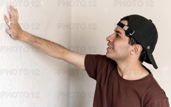 A young man inspects a wall closely during a home remodeling project, poised to begin his next task. Wearing a casual outfit and a baseball cap, he demonstrates a focus typical of DIY home improvement efforts