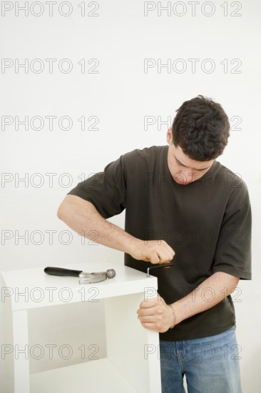 A young man in a casual t-shirt and jeans focuses intently on assembling a new white furniture shelf using a screwdriver and hammer