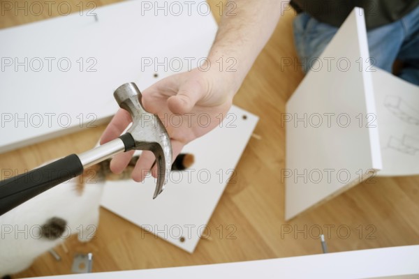 Cropped view of a man holding a hammer while assembling new furniture. The image shows his hands, the tool, and parts of the furniture on a wooden floor