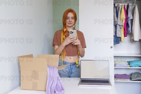 A young woman organizes clothes for online resale, using a laptop and smartphone. A box is ready for packing Her tidy closet is seen in the background, indicating efficiency