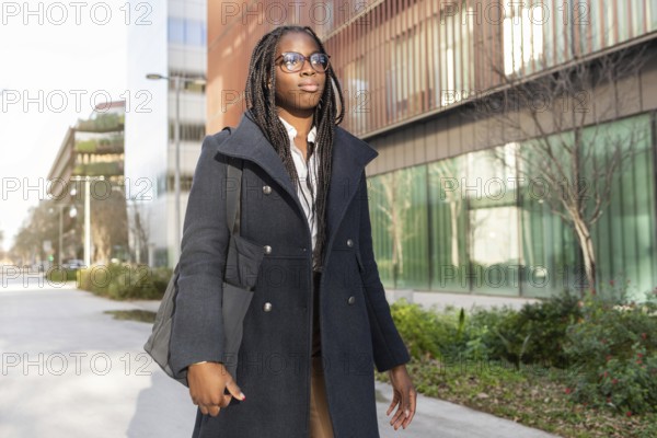 African American businesswoman with braided hair dressed in a stylish coat, takes a break outdoors in a modern city environment