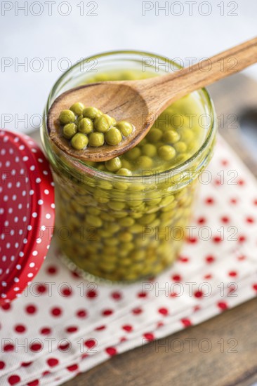 Close up of vibrant green homemade organic peas in a glass jar, with a wooden spoon resting on top. The jar sits on a white cloth with red polka dots, next to a matching lid