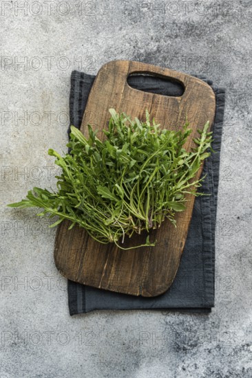 Bunch of fresh arugula placed on a rustic wooden cutting board with a dark cloth, set against a textured stone background. Perfect for natural and healthy food concepts