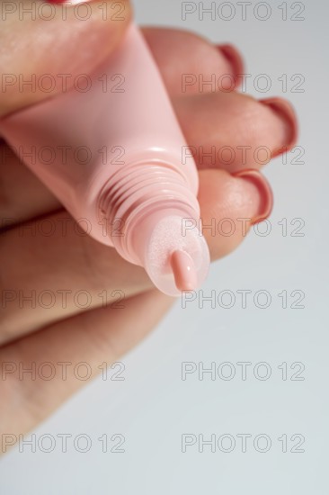 Cropped unrecognizable woman's hand applying a shiny lip balm from a pink tube, showcasing cosmetic textures on the skin in macro detail