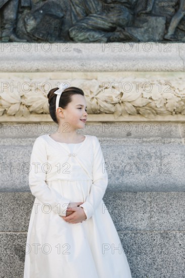A young girl stands in a beautiful First Communion dress, framed by an ornate stone wall with intricate carvings, capturing the essence of a cherished religious milestone