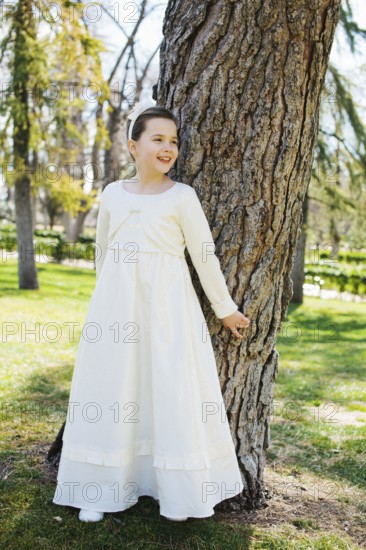 A young girl stands by a tree, smiling in a beautiful white First Communion dress. Sunlight filters through the leaves, creating a serene and joyful atmosphere