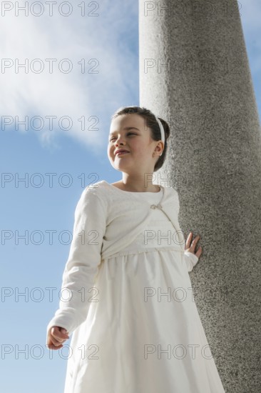 A young girl wearing a white First Communion dress stands outdoors against a stone column. The clear blue sky and clouds create a serene and joyous atmosphere