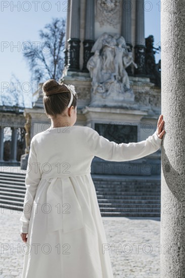 A young girl wearing a white First Communion dress stands beside a stone column, with a historic monument in the background, under a clear blue sky