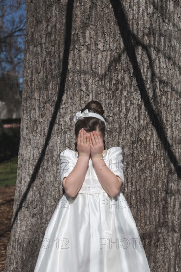 A young girl stands in front of a tree, covering her face with her hands. She is wearing an elegant First Communion dress, symbolizing a special religious occasion