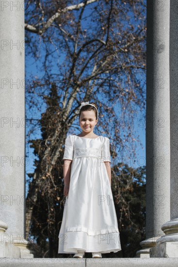 A young girl stands in a white First Communion dress, framed by stone columns and bare trees, with a bright blue sky providing a vivid backdrop to this special occasion