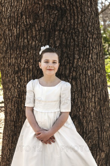A young girl in a First Communion dress stands gracefully against a tree, embodying innocence and joy. Her serene expression highlights the significance of the occasion