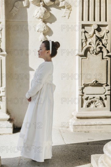 A young girl wearing a First Communion dress stands by an ornate wall. The scene captures a serene moment, highlighting her elegance and the architectural beauty around her