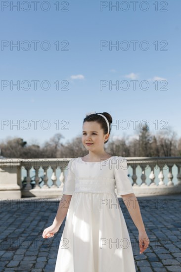 A young girl enjoys a sunny day outdoors, dressed in a beautiful white First Communion dress. The bright blue sky forms a picturesque backdrop to this serene moment
