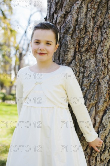 A young girl stands by a tree, wearing a white First Communion Day dress. She smiles softly under the sun, surrounded by the serene ambiance of a lush park