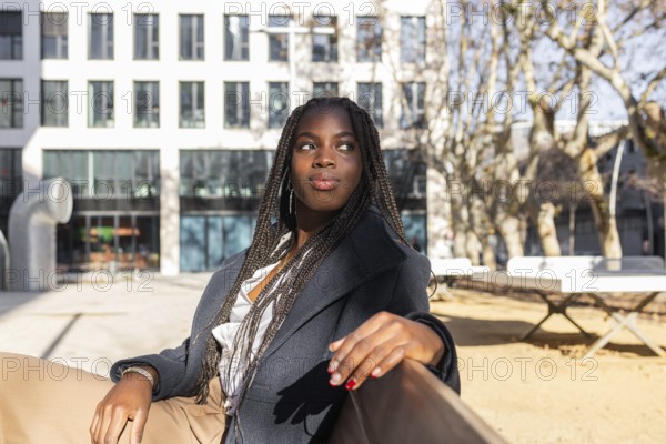 A young African American businesswoman with braids takes a relaxing break outdoors, sitting with a confident look against an urban backdrop