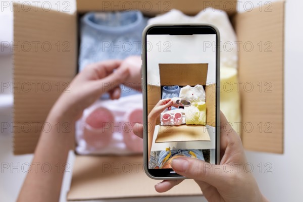 A young woman uses her smartphone to photograph clothing items for an online sale. She holds the phone while organizing clothes in a box, capturing each piece for listing