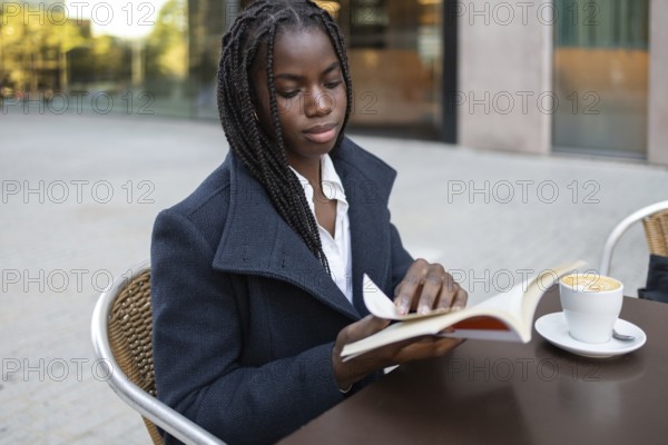 An African American businesswoman with braids is sitting on an outdoor cafe table during a break, reading a book while sitting by a coffee cup
