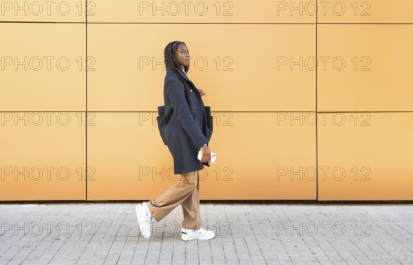 A young African American businesswoman with braids, dressed in a formal grey jacket and beige trousers, holding a smartphone while walking against a vibrant yellow wall