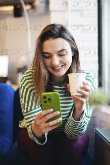 A young woman in a striped green and white top happily operates her smartphone while enjoying a cup of coffee in a cozy cafe setting