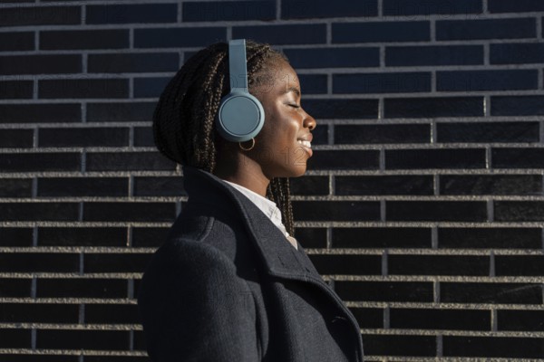 African American businesswoman with braids smiles while listening to music with headphones during a break outdoors, against a brick background