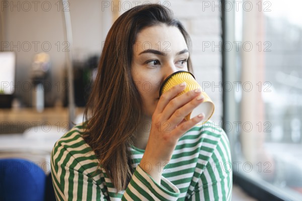A young woman in a striped shirt takes a peaceful coffee break, sipping from a yellow cup inside a modern cafe, showcasing a relaxed and contemplative moment