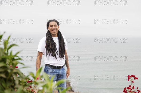 A man with long dreadlocks and tattoos smiles warmly, standing near vibrant flowers with the serene ocean as a backdrop in Barranco, Lima, Peru A scene of natural beauty and culture
