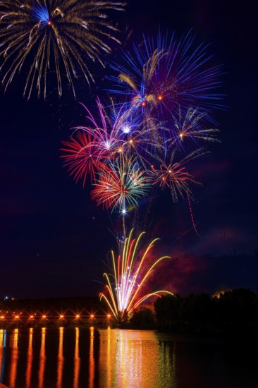 Vibrant fireworks illuminate the night sky above the Ebro River, reflecting on its calm waters. The image captures a spectacular array of colors and patterns during a fireworks event