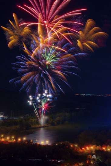 Vibrant fireworks illuminate the night sky above the Ebro River, casting colorful reflections on the water, surrounded by a dark landscape lit by streetlights