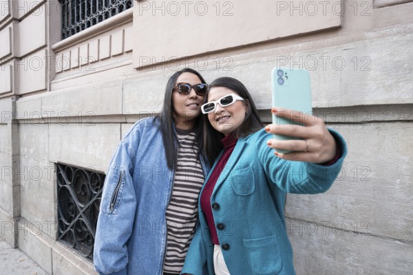 A Latin lesbian couple takes a joyful selfie outdoors, leaning against a historic building. Both wear stylish jackets and sunglasses, capturing a moment of love and happiness