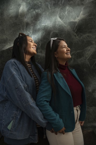A joyful Latin lesbian couple shares a heartfelt moment, smiling warmly. They are dressed stylishly, with a textured dark background highlighting their happiness