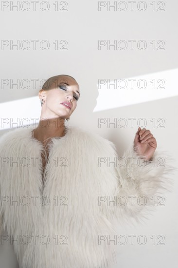 A confident middle-aged woman with a shaved head poses elegantly in a luxurious faux fur coat. Minimalist lighting highlights her serene expression and sophisticated style