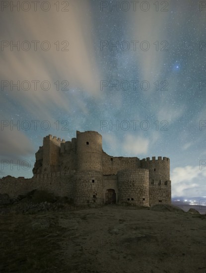 The medieval Manqueospese Castle in Avila, Spain, stands under a starry sky, with ethereal clouds drifting over its ancient stone walls
