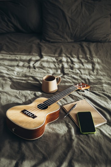 A cozy and creative setting featuring a ukulele, a ceramic mug, an open notebook with a pen, and a smart phone, all placed on a soft, wrinkled bedspread