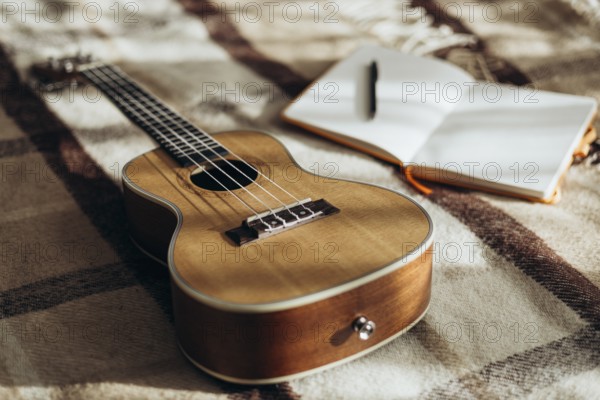A ukulele rests on a cozy, plaid blanket beside an open notebook with a pen, capturing a serene, creative atmosphere with natural lighting, perfect for music and writing