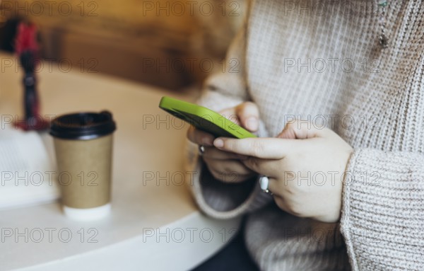 Close-up of a woman's hands holding a green smartphone in a bookstore cafe A takeaway coffee cup and books on the table suggest a relaxed and focused moment in a cozy space