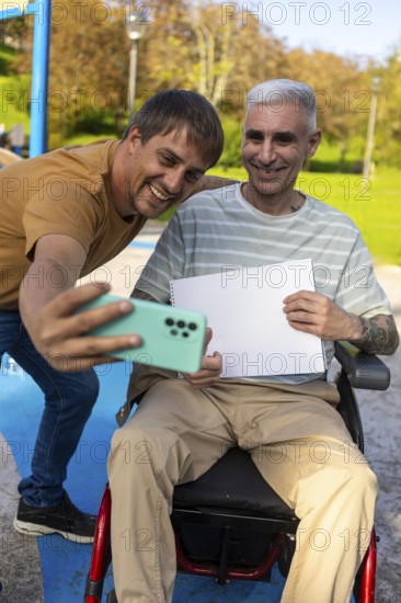 Two friends in a park enjoy taking a selfie together One person is sitting in a wheelchair by cerebral palsy, holding a blank notebook page, while the other captures the moment