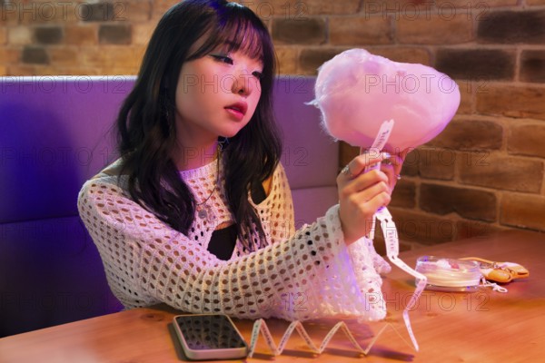 Young Asian Gen Z girl enjoys a vibrant arcade environment, sitting at a colorful booth with cotton candy and tickets, capturing the essence of youthful leisure and joy